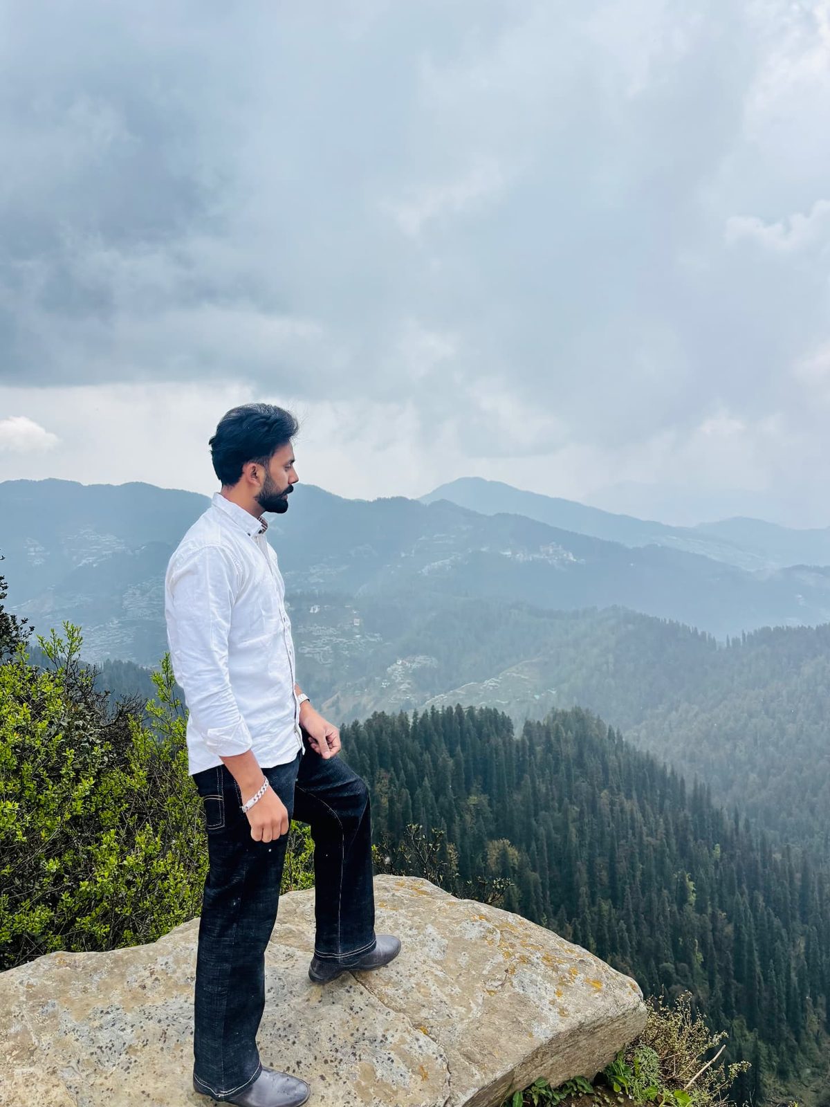 Sanjam standing on a rock at a cliff edge overlooking pine-covered hills under grey clouds.