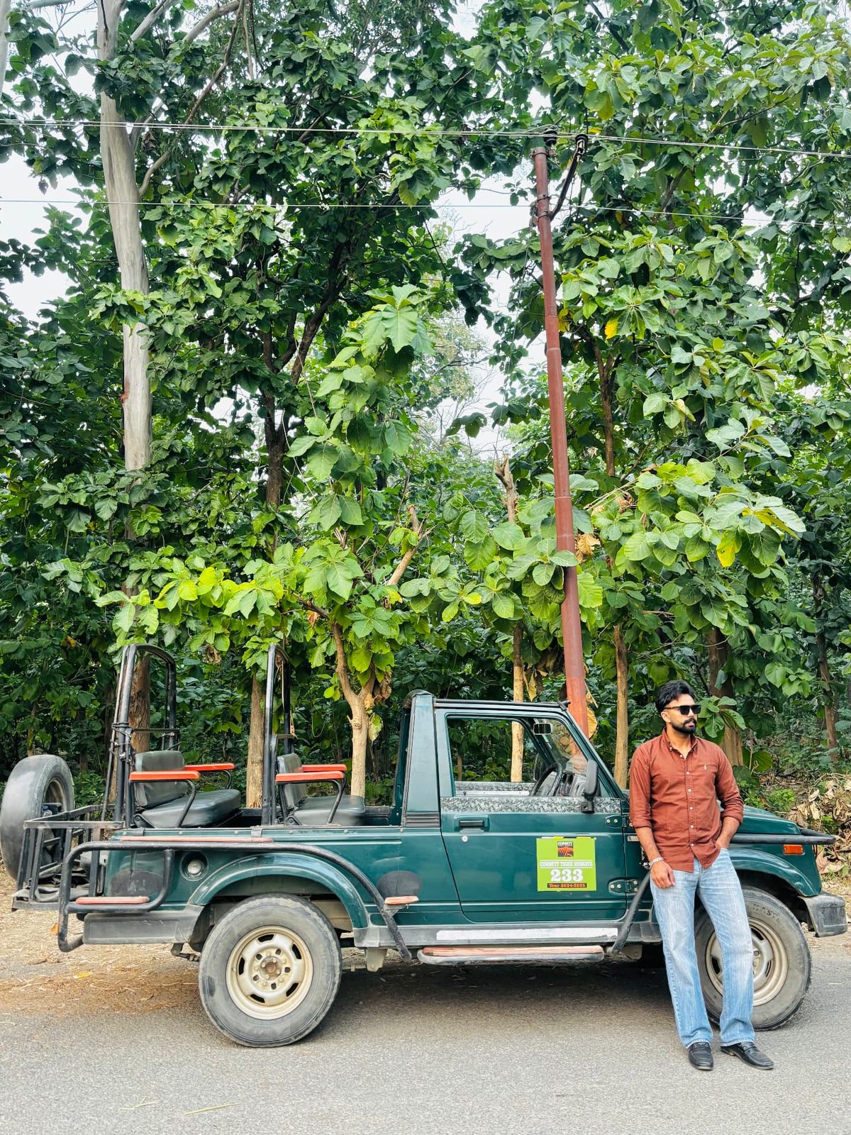 Sanjam leaning against a green open-top safari jeep parked under tall green trees.