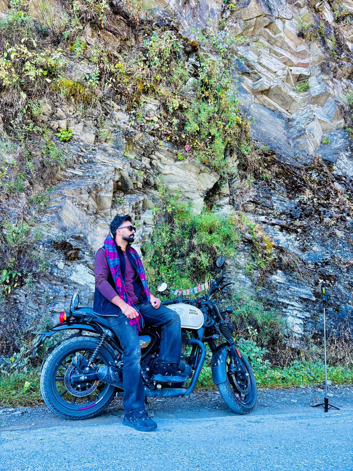 Sanjam sitting on a black Royal Enfield motorcycle beside a rocky mountain roadside.
