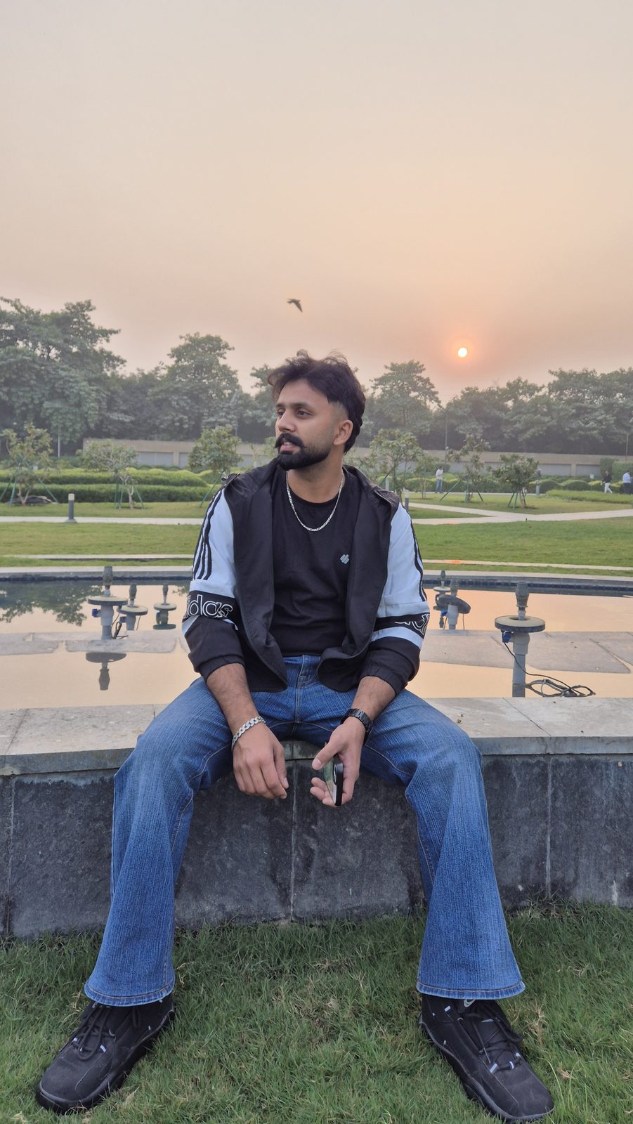 Sanjam sitting on a stone ledge by a reflecting pool as the sun sets behind trees.