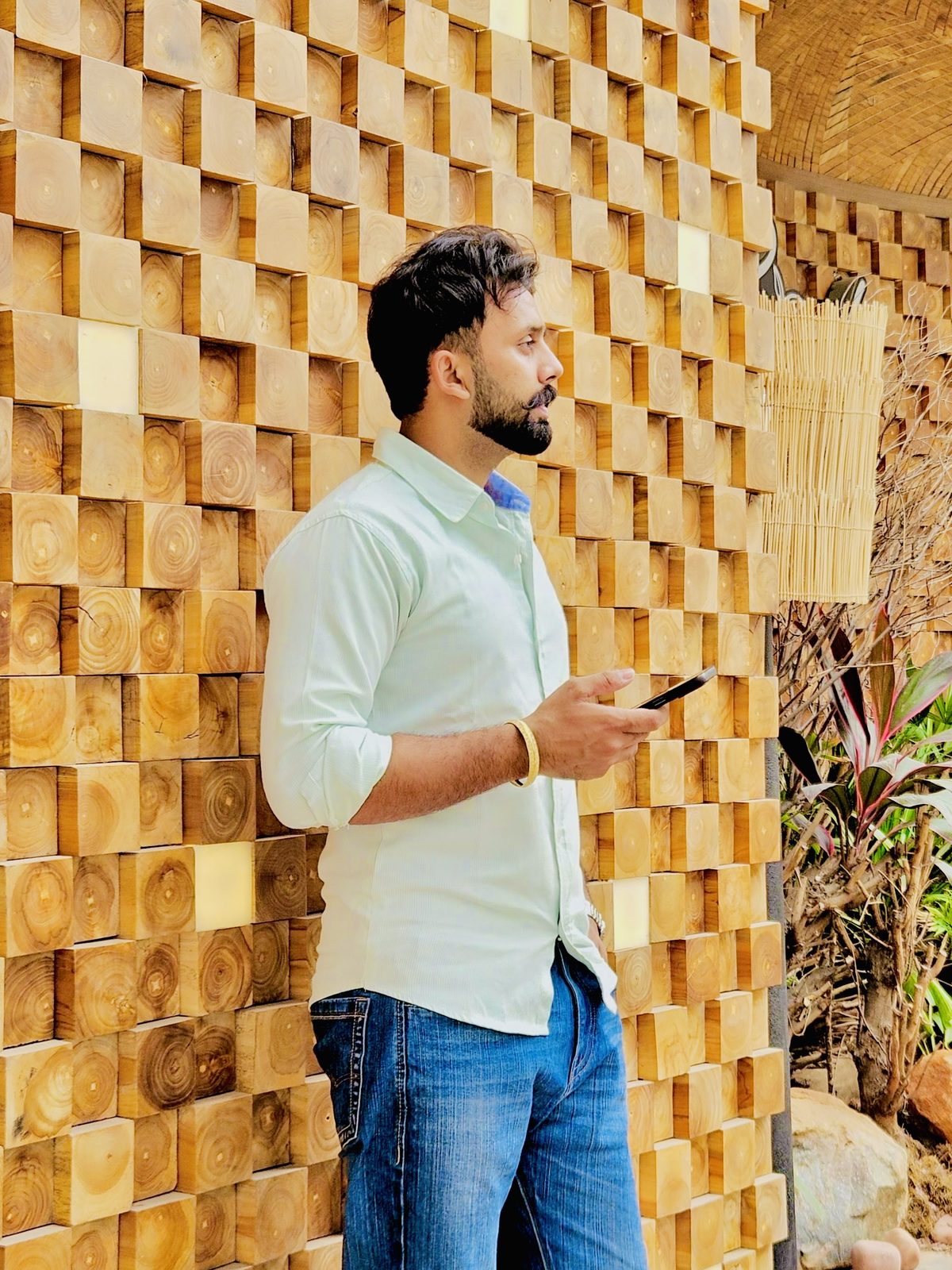 Sanjam in a mint shirt standing in front of a wall of stacked teak wood blocks.